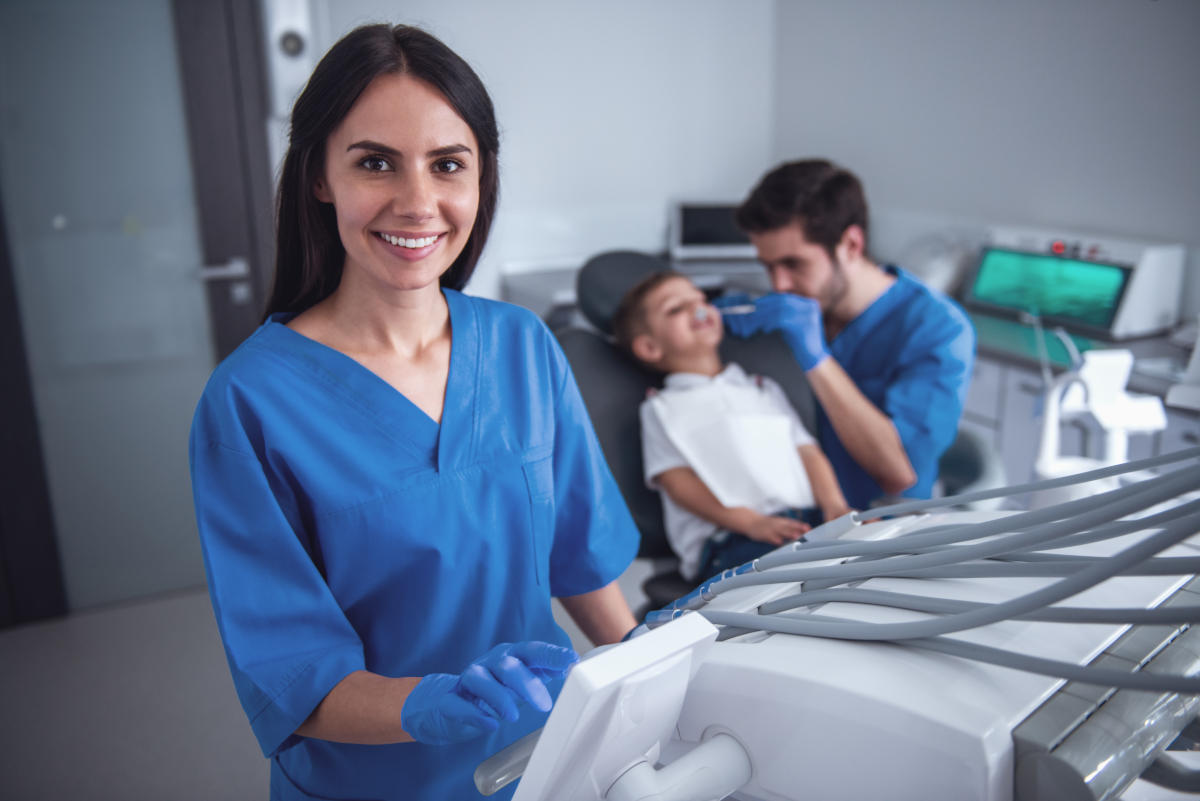 Beautiful young female dentist is looking at camera and smiling, little boy visiting dentist in the background